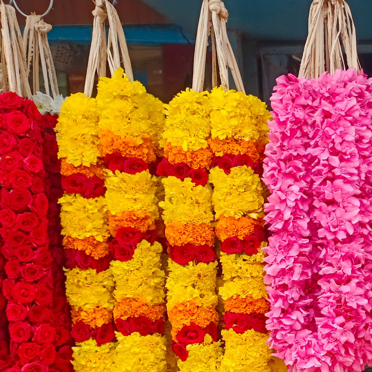 Yellow chrysanthemum garlands banded with orange and red, between a red rose garland and a pink garland, at a market stall