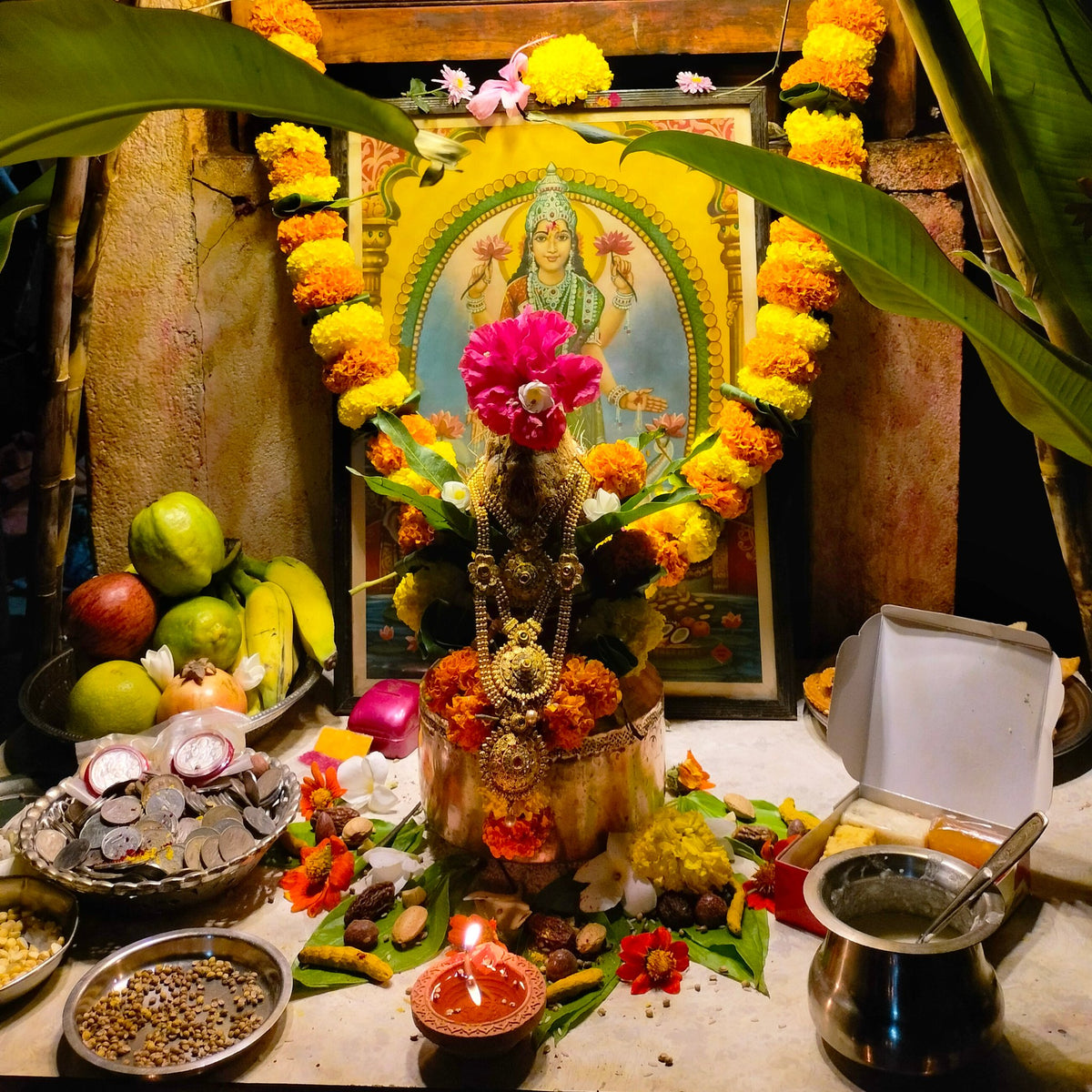 A Lakshmi pooja set up for Diwali with marigold garlands, a decorated kalasam, fruit and a lit diya