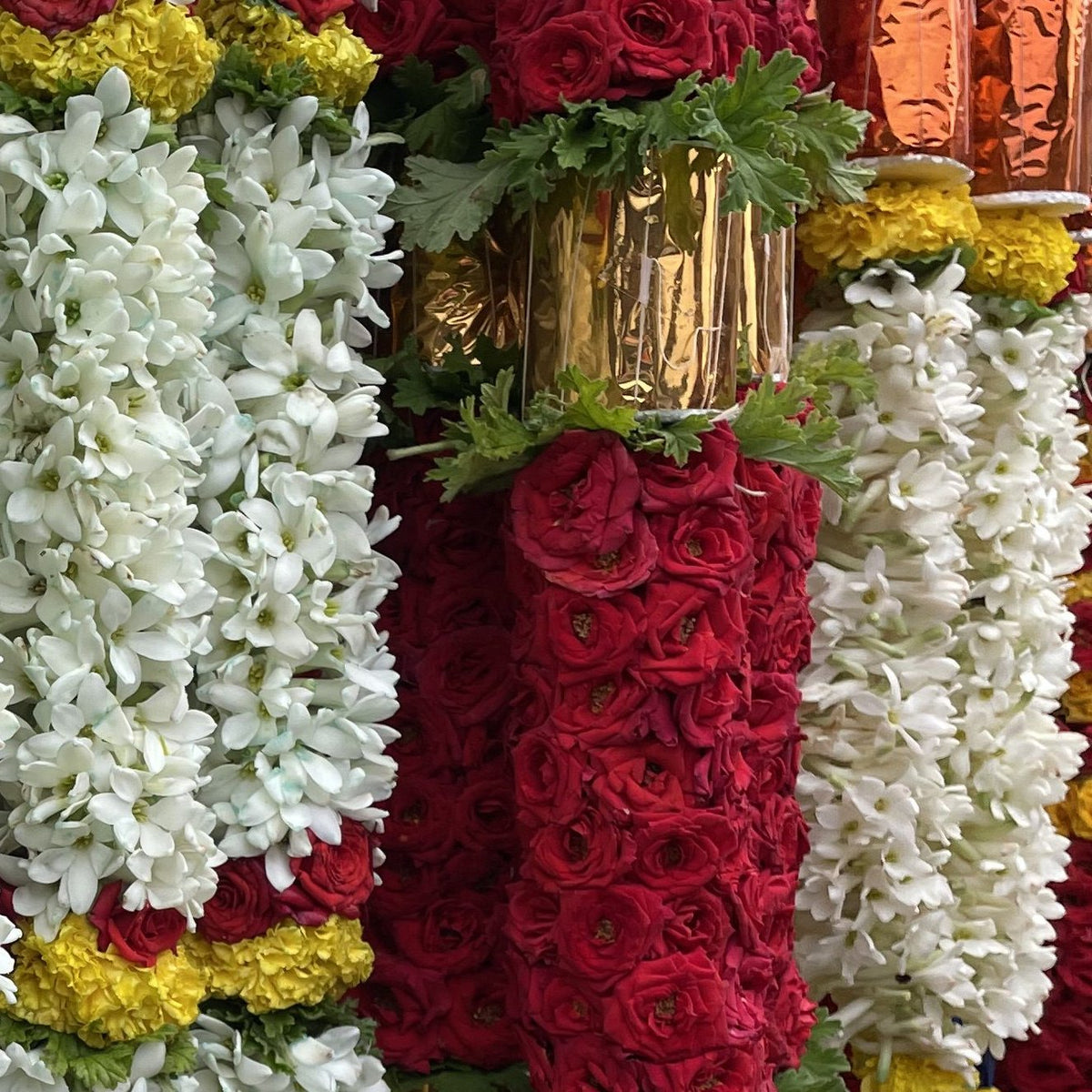 A thick red rose garland with a gold foil band and green leaves, between white tuberose garlands