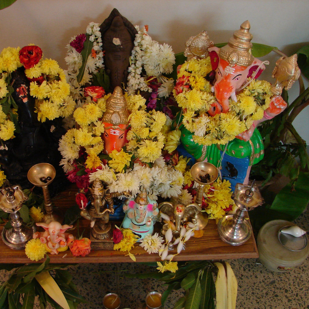 A home Ganesh Chaturthi altar with chrysanthemum and jasmine garlands, mango and banana leaves and brass lamps