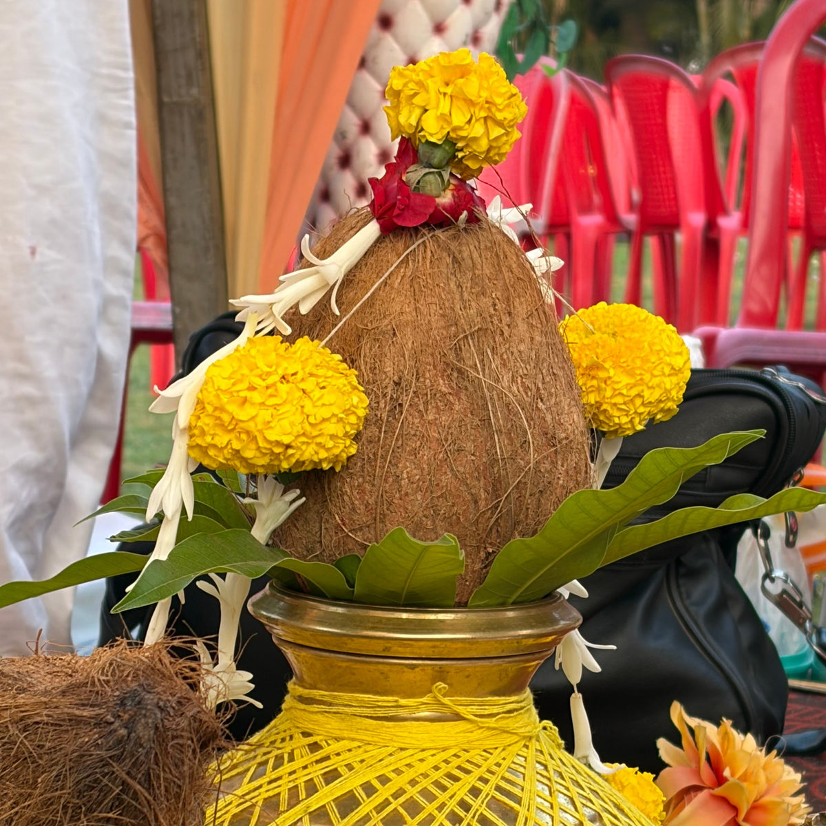 A coconut on a brass kalash wrapped in yellow thread, decorated with marigolds, a red rose, tuberose strands and mango leaves