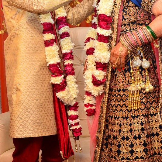A bride and groom, seen from the shoulders down, wearing garlands of white chrysanthemum and red rose